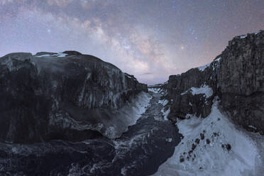 Panoramic view of a frozen river flowing through a snow-covered canyon with sheer cliffs, all under a dim evening starry sky in Iceland - ADSF49681