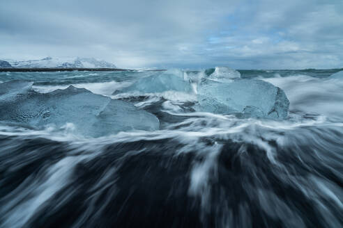 Picturesque scenery of rippling sea water with pieces of broken ice on black sandy beach against cloudy sky in Diamond Beach in Iceland - ADSF49689