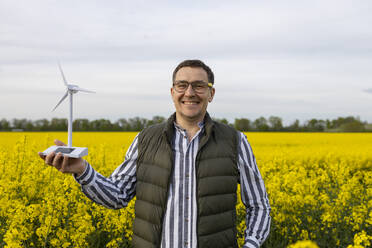 A smiling mature man with glasses stands in a vast yellow flower field, presenting a model of a wind turbine. - ADSF49690