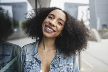 Joyful afro young woman with curly hair wearing a denim jacket and white top smiling and looking at camera while leaning against a reflective glass pane against blurred city - ADSF49692