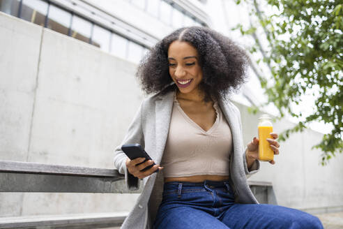 Happy ethnic young woman with curly hair sitting outdoors near a modern building and holding a smartphone in one hand and a small bottle of juice in the other - ADSF49694