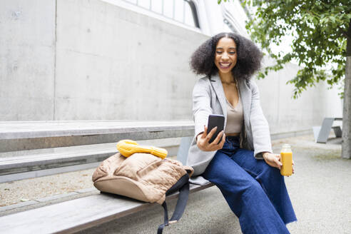 Happy ethnic young woman with curly hair sitting outdoors near a modern building and taking selfie with smartphone while holding a small bottle of juice - ADSF49696