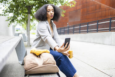 Happy ethnic young woman with curly hair sitting outdoors near a modern building and taking selfie with smartphone while holding a small bottle of juice - ADSF49697