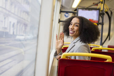 Joyful ethnic woman with curly hair waving hello and looking at camera while sitting on bus seat - ADSF49699