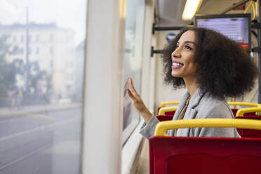 Joyful ethnic woman with curly hair gazes out the window of a city bus while admiring the outside view in street - ADSF49700