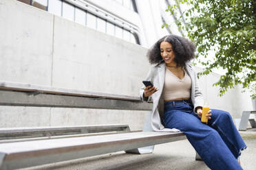 Happy ethnic young woman with curly hair sitting outdoors near a modern building and holding a smartphone in one hand and a small bottle of juice in the other - ADSF49701