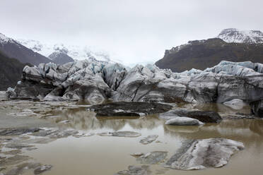 Jagged ice formations arise from a murky glacial lagoon surrounded by dark mountains under a clouded sky in Iceland - ADSF49703