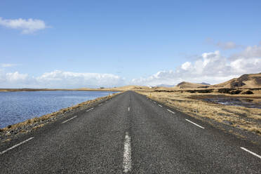 Diminishing perspective of asphalt empty roadway with road markings passing through scenic lake against sky in Iceland - ADSF49713