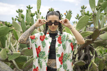 Modern young tattooed woman in casual clothes looking at camera while standing and adjusting sunglasses against blurred nature in Masca Tenerife, Canary Islands - ADSF49717