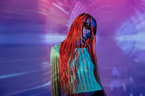 Portrait of emotionless young woman with orange braids wearing sunglasses and posing confidently against neon glowing lights - ADSF49722