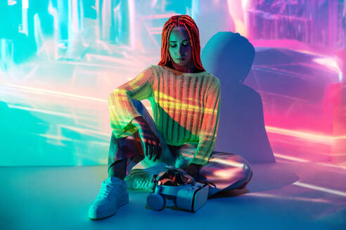 Full body of serious thoughtful young woman with modern VR goggles sitting in glowing lights while exploring cyberspace in studio - ADSF49723