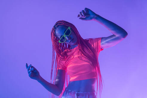 Portrait of emotionless young woman with orange braids wearing sunglasses and posing looking down confidently against neon purple glowing lights - ADSF49726