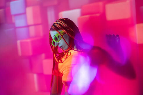 Portrait of emotionless young woman with orange braids wearing sunglasses and posing looking down confidently against neon pink glowing lights - ADSF49727