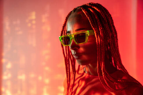 Side view portrait of emotionless young woman with orange braids wearing sunglasses and posing looking down confidently against neon orange glowing lights - ADSF49728