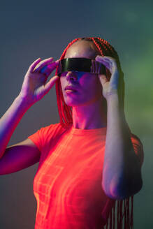 Beautiful woman with orange braids in trendy VR glasses experiencing virtual reality against colorful background - ADSF49729