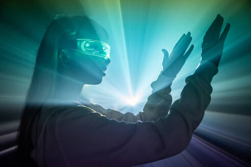 Side view of young woman wearing futuristic LED VR glasses moving arms projected lights while exploring cyberspace in studio - ADSF49732