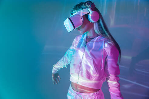 Unrecognizable young woman in casual outfit watching through modern VR goggles and immersed in exploration of cyberspace on projected glowing lights - ADSF49733