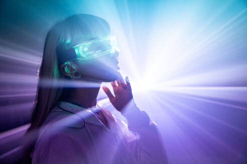 Side view of unemotional young woman wearing futuristic LED VR glasses touching face with projected lights while exploring cyberspace in studio - ADSF49734