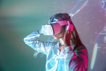 Side view of unrecognizable young woman in casual outfit watching through modern VR goggles and immersed in exploration of cyberspace on projected glowing lights - ADSF49735