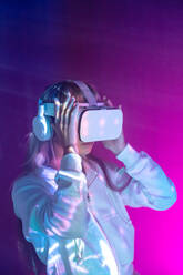 Unrecognizable young woman in casual outfit watching through modern VR goggles and immersed in exploration of cyberspace on projected glowing lights in purple background - ADSF49736