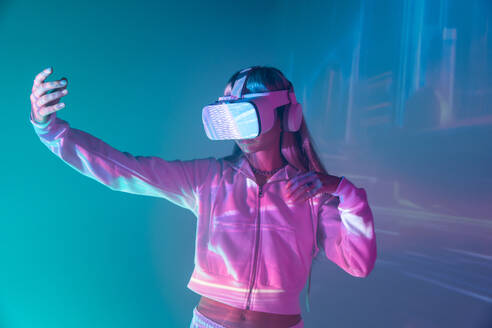 Unrecognizable young woman in casual outfit watching through modern VR goggles and moving hands in immersed exploration of cyberspace on projected glowing lights - ADSF49737