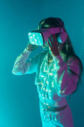 Side view of unrecognizable young woman in casual outfit watching through modern VR goggles and immersed in exploration of cyberspace on projected glowing lights - ADSF49738