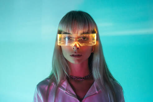 Portrait of confident unemotional young woman wearing LED futuristic VR glasses and looking at camera in blue background - ADSF49739