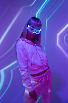 Serious unemotional young woman in casual attire with eyes closed experiencing virtual reality through modern LED VR goggles in studio with glowing projected lights against purple background - ADSF49740