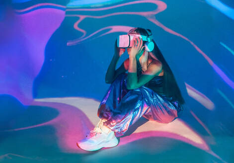 Side view of unrecognizable young woman in casual outfit watching through modern VR goggles and immersed in exploration of cyberspace on projected glowing lights while sitting on floor - ADSF49743