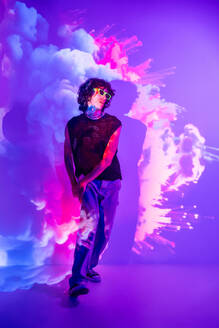 A person stands amidst vibrant clouds of pink and white smoke, highlighted by purple lighting, wearing sunglasses and a mesh top - ADSF49752