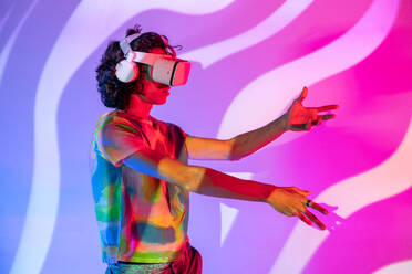 Young man wearing a tie-dye shirt and VR headset reaches out, immersed in the digital world, complemented by a wavy pink and purple background - ADSF49756