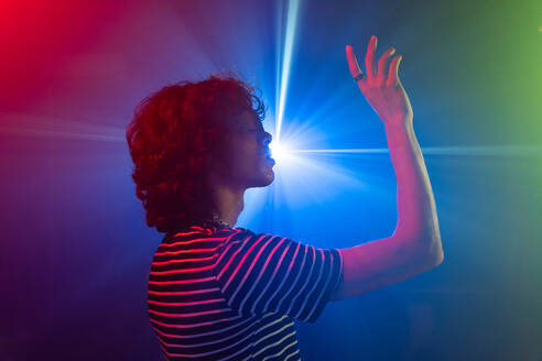 Curly-haired man and futuristic glasses in a striped shirt reaches towards a radiant blue light, surrounded by a moody red and purple ambiance - ADSF49757