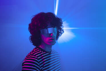 Young man with curly hair and a striped shirt wears futuristic glasses, illuminated by a piercing blue light in a deep purple setting - ADSF49758