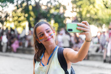 Young woman taking a selfie outdoors during a pride event with face adorned with colored stripes symbolizing LGBTQ+ pride against blurred people in background - ADSF49762