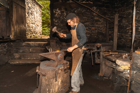 Side view of skilled blacksmith adorned in worn clothing meticulously hammers a heated piece of metal atop an anvil shaping a distinctive 18th century hydraulic mallet inside a dimly lit workshop - ADSF49766