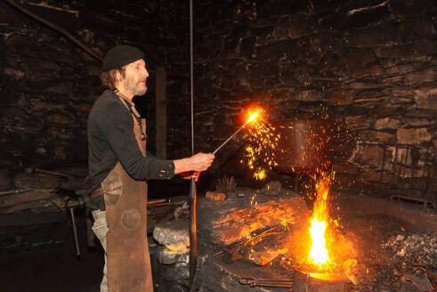 A focused blacksmith in traditional attire stands by a blazing forge with sparks flying as he works on forging an 18th century hydraulic mallet emphasizing the rarity of the craft in Europe - ADSF49767