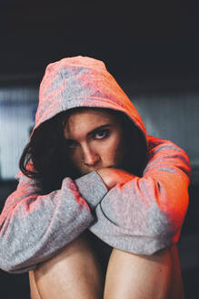 Scared woman wearing hooded shirt and sitting in garage - SVCF00414