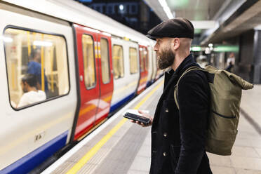 Pendler mit Smartphone in der Hand, der am Bahnhof auf seinen Zug wartet - WPEF07876