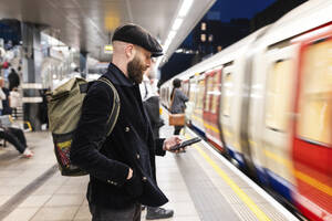 Mann mit Smartphone in der Nähe eines fahrenden Zuges am Bahnhof stehend - WPEF07877