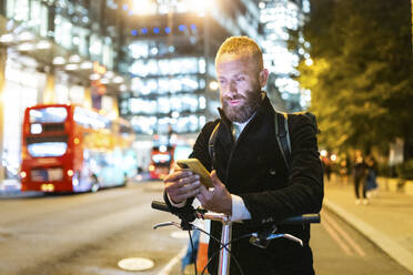 Pendler mit Fahrrad und Smartphone am Straßenrand in der Stadt - WPEF07881