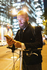 Man using smart phone leaning on cycle in city at night - WPEF07882