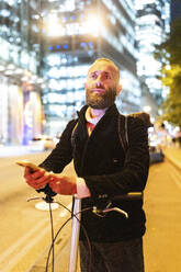 Man holding smart phone and standing with cycle in city at night - WPEF07883