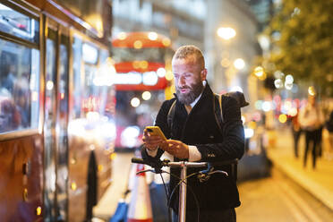 Mann mit Mobiltelefon, stehend mit Fahrrad in der Stadt bei Nacht - WPEF07885