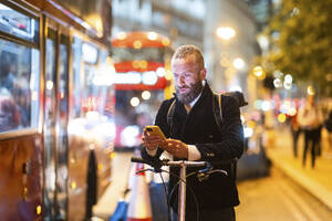 Mann mit Mobiltelefon, stehend mit Fahrrad in der Stadt bei Nacht - WPEF07885