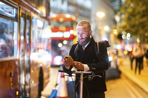 Man using mobile phone standing with cycle in city at night - WPEF07885