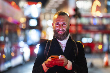 Smiling mature man holding smart phone in city at night - WPEF07895