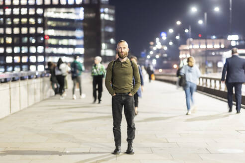 Man with hands in pockets standing on footpath in city at night - WPEF07905