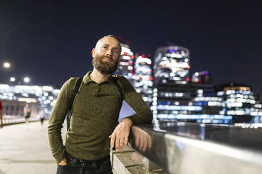 Man with hand in pocket leaning on railing in city at night - WPEF07908