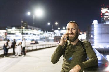 Smiling man talking on smart phone near railing in city - WPEF07910