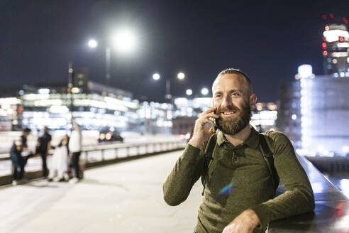Lächelnder Mann, der in der Nähe eines Geländers in der Stadt mit seinem Smartphone telefoniert - WPEF07910